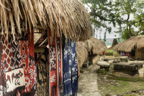 Woven ikat with natural dyes is displayed in front of a traditional Sumba house made of wood, bamboo, and a thatched roof. 
