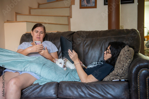 Mujer madura, latina Mexicana con su hija en un sofa revisando tareas con celuklar y tablet con la mascota acostada entre ellas, en la sala de su hogar, con escaleras a segundo piso en el fondo