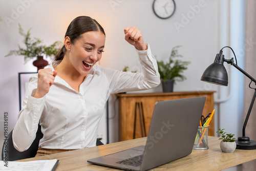 Young businesswoman at home office views growth chart on laptop sees profit jump and smiles raising hands. Freelancer girl at table celebrates success says yes aloud shares win by message to client.