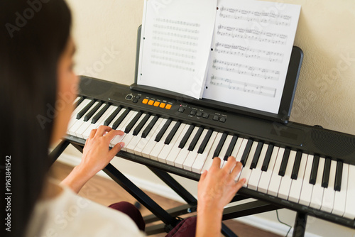 Young woman learning to play keyboard, practicing digital piano with sheet music in home setting