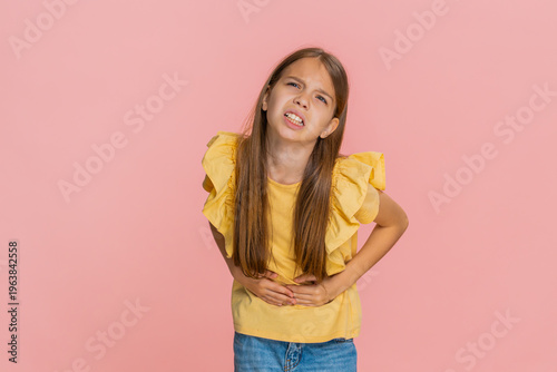 Young child girl holds her stomach with frightened expression, showing that something hurts and feeling unwell. School kid isolated on pink background showing pain and reaction related to health issue