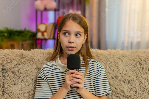 Young child girl shyly sings into a microphone, smiling quietly while sitting on home sofa and enjoying the moment. School kid performs alone, expressing gentle emotions through soft voice sweet charm
