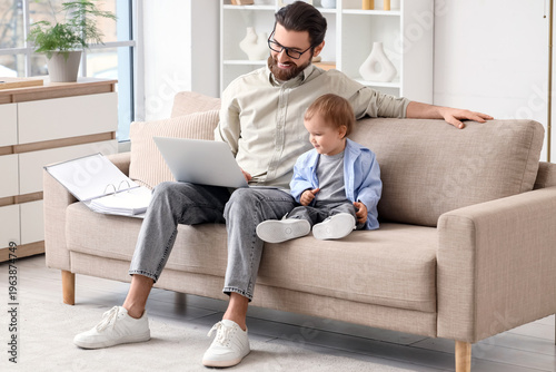 Father working with laptop and his little baby on sofa during parental leave ...