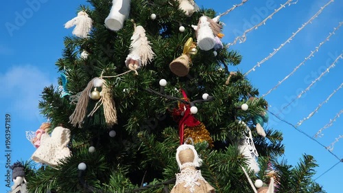 Colorful ornaments, shimmering lights, and cheerful angels create a festive scene on a majestic tree. The bright blue sky enhances the joyful winter atmosphere, inviting all to celebrate