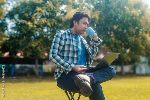 Εκτύπωση καμβά A person is sitting on a folding chair in a park, holding a laptop and a cup, dr