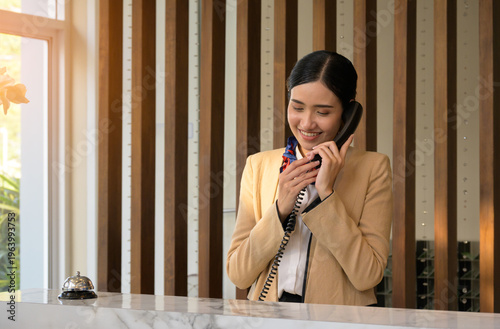 Smiling Asian female hotel receptionist in yellow blazer talking on landline telephone at luxury hotel front desk with service bell