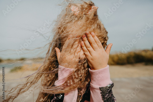 The little girl covers her eyes with her hands, the wind blowing her long hair.