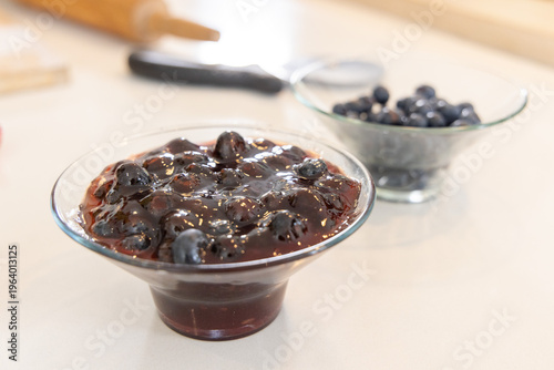 Glass bowl of blueberry jam sitting on counter with blueberries, rolling pin blurred