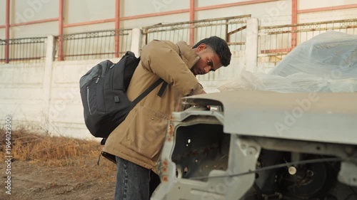 Technician examining vehicle in abandoned lot. Worker repairing vehicle engine amidst scrapyard in autumn daylight. Man inspects car engine carefully while dressed for colder weather in dirt yard