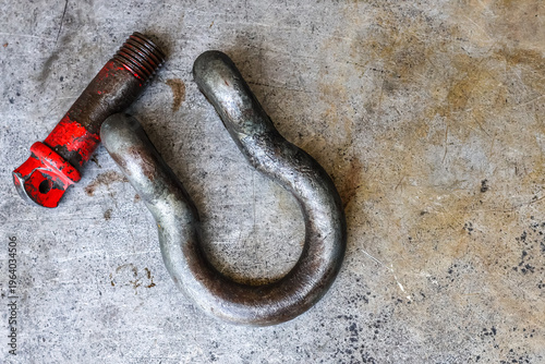 Foto ​A heavy-duty, worn, and rusted industrial shackle and a detached red-painted screw pin rest side-by-side on a scratched metal workshop floor, capturing detailed texture and a rugged feel