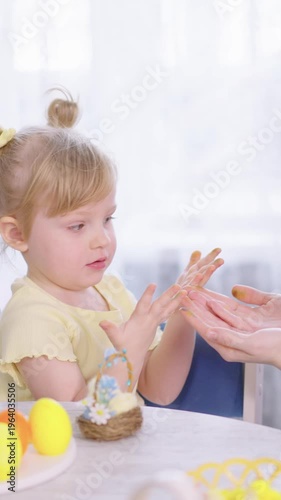 Little girl smiles, delighted by colorful Easter eggs. She claps, joyful, as someone helps her with craft. Soft light and playful table scene capture happy family moment