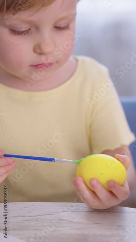 Child paints a yellow ball with care. Focused eyes show quiet concentration. Soft light highlights the gentle task. Perfect for creative or parenting themes. No animals or plants present in this scene