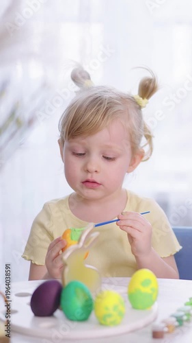 Young girl paints Easter eggs with care. Her focused gaze shows quiet concentration. She holds a brush, dips it in dye. Colorful eggs rest on a white plate. Bright, soft light fills the room