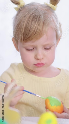 Soft light illuminates a child's focused face. Tiny hands steady paintbrushes over colorful eggs. Child's eyes show quiet concentration and delight. Perfect for Easter, parenting
