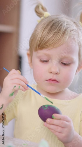Child paints an egg with care and focus. Soft light highlights her concentration. Yellow dress matches her cheerful energy. Eyes show quiet determination. Perfect for spring or family activity scenes