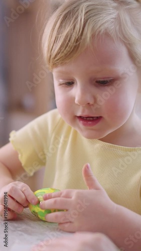 Soft light highlights a young child's focused expression. Tiny hands explore a colorful egg-shaped toy. Child's gaze follows movement with quiet curiosity