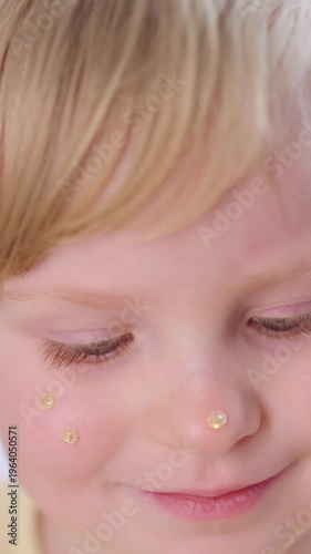 Little girl smiles while applying golden dots to her cheeks. Soft light highlights her playful expression. Curly blonde hair frames her gentle face. The joyful moment captures innocence and fun