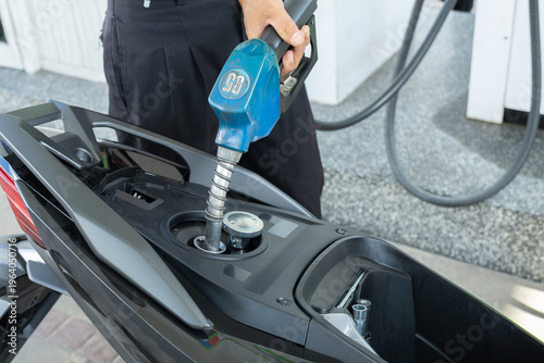 A hand holds a blue nozzle, filling 95 RON gasoline into a black scooter at a Thai gas station, capturing everyday fuel use.