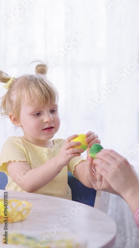 Soft light fills a cozy home setting. Child holds a painted egg, eyes wide with wonder. Adult gently offers a second colorful egg. Warm interaction highlights bonding moments
