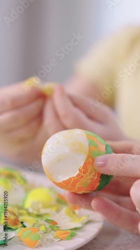 Close-up of hands peeling a painted egg. Soft light illuminates the gentle activity. Eggshell fragments scatter on a plate nearby. Child's hands join in the creative process
