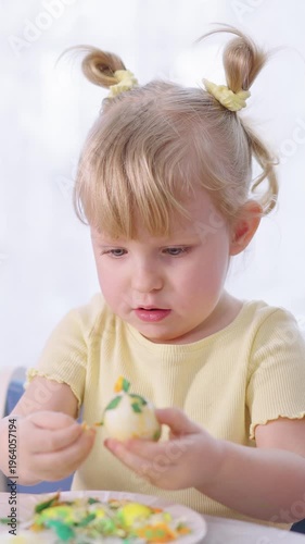 Little girl peels a hard-boiled egg. Curious eyes focus on the shell. Hands work carefully to crack the egg. Soft yellow shirt matches her hair. Bright background adds warmth to scene