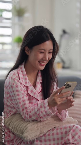 Asian adult woman smiling enjoying mobile phone content sitting sofa cozy living room pajamas browsing digital media online for relaxation