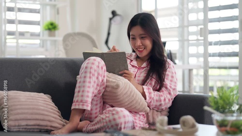 Young Asian woman leisure reading writing personal journal comfy pajamas sofa home living room wellness relaxation calm