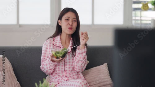 Asian adult woman eats healthy fresh salad in comfortable pajamas relaxing on home sofa watching television enjoying wellness lifestyle