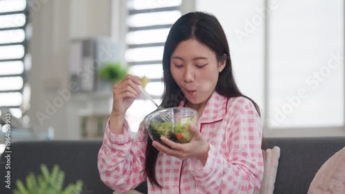 Asian woman enjoys healthy salad meal wearing pajamas at home sofa adult female relaxed eating fresh green diet nutrition wellness lifestyle