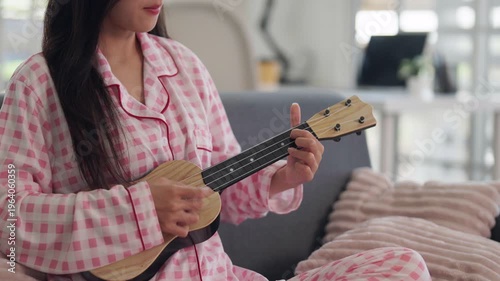Asian young adult woman plays ukulele relaxing at home living room comfort music hobby leisure time activity