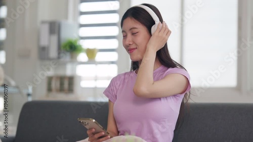 Asian young adult woman enjoying peaceful audio content using smartphone and headphones relaxing comfortably in modern home living room