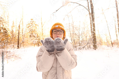 A boy blows snow from her mittened hands in a sun-drenched winter forest, eyes closed, breath visible, radiating cozy joy and childlike wonder.