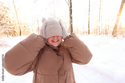 A girl grins playfully while hiding her eyes behind a knit beanie and mittens in a sunlit snowy forest, joyful, mischievous, and wrapped in winter warmth.