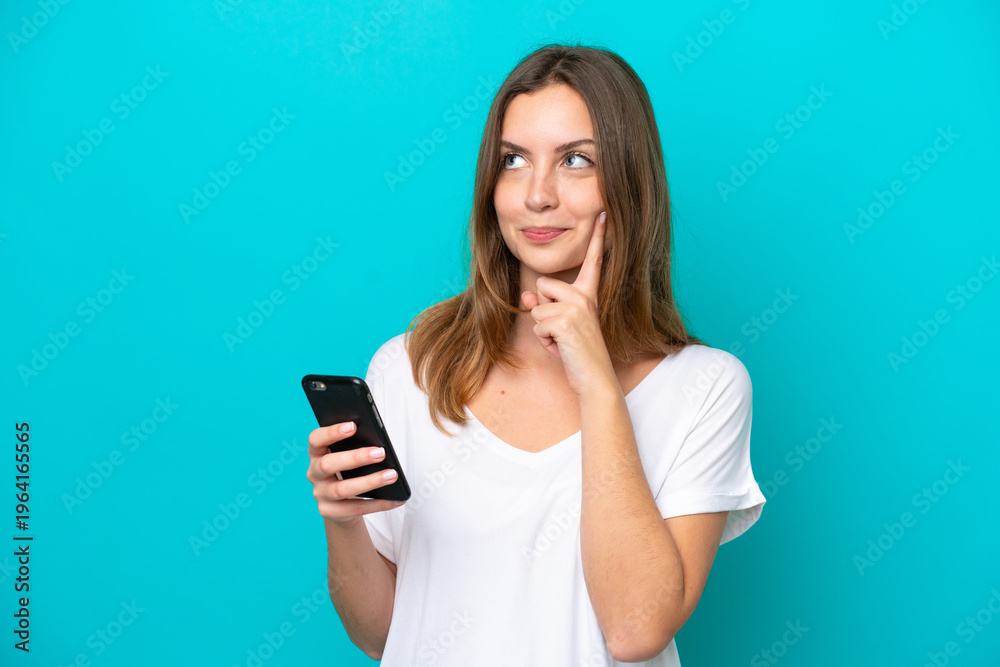 Fototapeta premium Young caucasian woman isolated on blue background using mobile phone and thinking