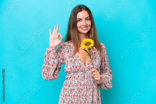 Young caucasian woman holding sunflower isolated on blue background showing ok sign with fingers