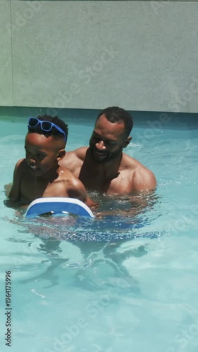Vertical video: Guiding dad holding kickboard, teaching son in trunks kicking in pool, green ring
