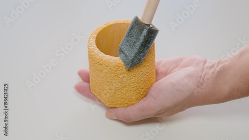 Hand Holding Yellow Textured Planter Pot With Green Succulent Leaf Against White Background