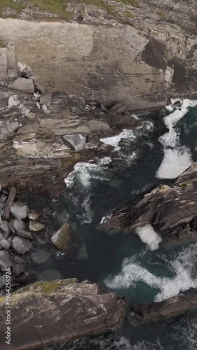 Powerful ocean waves break against dark, jagged rock formations and cliffs. White foam contrasts with the deep blue-green water, highlighting the natural power of the sea at this rugged coastline.