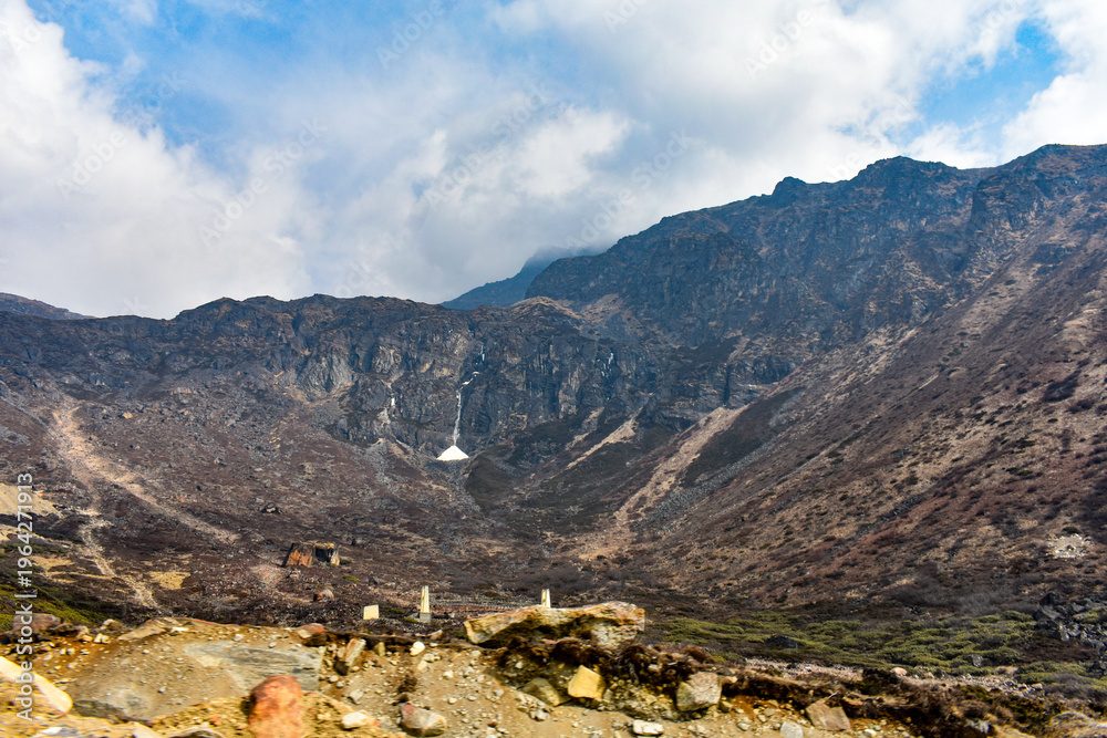 Fototapeta premium Rocky Himalayan valley with steep cliffs, waterfall, and rugged terrain under cloudy sky in North Sikkim landscape.