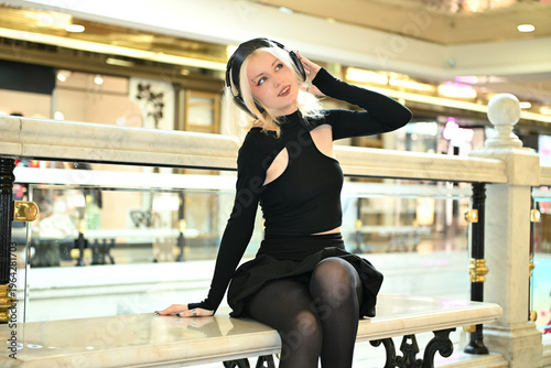 A young blonde girl of European appearance in a black dress listens to music through wireless headphones