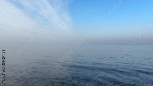 perfectly calm, glassy sea beneath a vast pale blue sky with wispy clouds, where the horizon dissolves into soft coastal mist, creating an exceptionally minimal and serene seascape