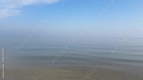 perfectly calm, glassy sea beneath a vast pale blue sky with wispy clouds, where the horizon dissolves into soft coastal mist, creating an exceptionally minimal and serene seascape