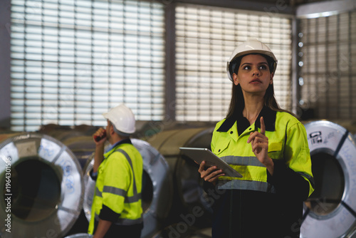 A woman worker wearing a yellow and blue safety vest is holding a tablet in a factory