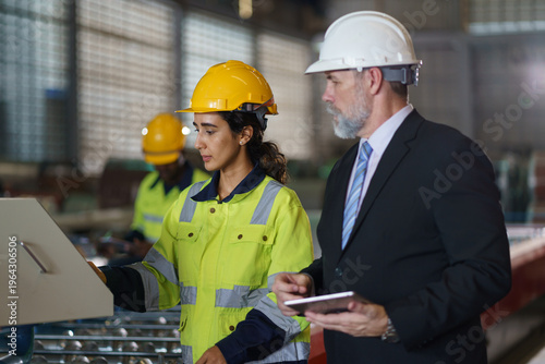 A foreman and a woman worker are standing in a factory, looking at a tablet. The engineer talks with the woman's workers. They wear a yellow and white safety helmet for safety.
