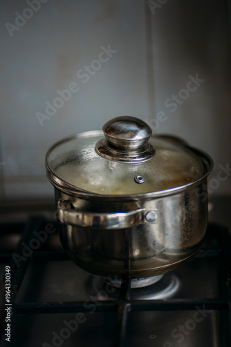 Close-up of a stainless steel cooking pot with a glass lid on a gas stove burner, steam condensing inside the lid during the boiling process, home kitchen cooking concept
