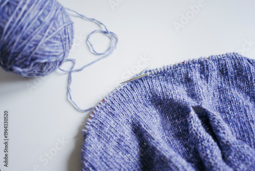 High-angle view of a skein of blue wool yarn and a knitting project on metal circular needles on a white background, cozy hobby and handmade craft concept