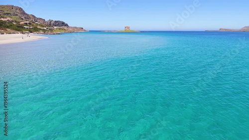 Low drone flight approaching the iconic ancient tower at La Pelosa beach in Sardinia. Clear turquoise water and white sand surround the historic landmark, creating a scenic Mediterranean coastal shot 