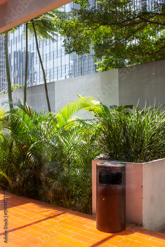 A black iron trash can stands on a red tile path next to a lush tropical garden with an office building background.