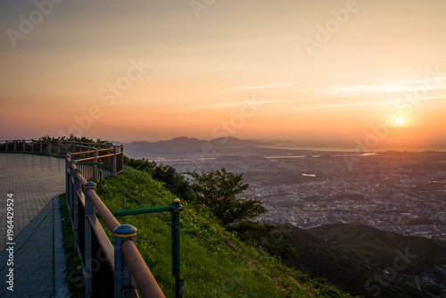 Panoramic sunset view from a mountain observatory overlooking the vast city lights and bay of Kitakyushu as the sun dips below the horizon.