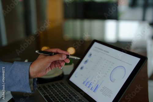 Businessperson's hand holding a pen while pointing at detailed financial charts and graphs displayed on a laptop screen, illustrating business analytics and data interpretation